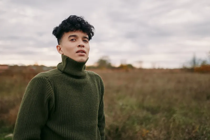 A genuine portrait of a young man standing in an open field, natural light highlighting his thoughtful expression and textured sweater, candid composition conveying realistic, trustworthy everyday