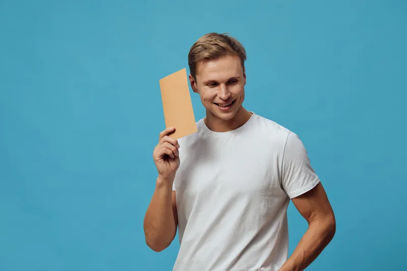 Smiling man holding a blank cardboard sign in front of a bright blue background. Casual style, cheerful expression, young adult, white t-shirt, studio shot, positive mood, advertising concept, face fr