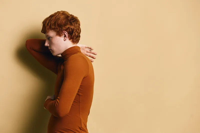 Redhead man with curly hair wearing brown turtleneck sweater posing sideways on beige background, thoughtful and relaxed expression with hand behind head in studio photo.