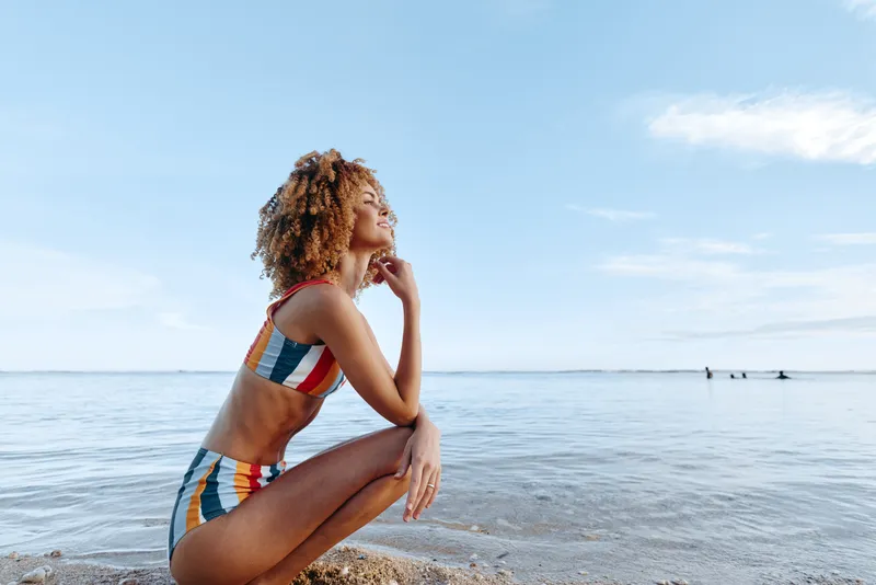 Vibrant beach scene with a woman in a striped bikini by the sea, sunlit moment, relaxed pose, joyful expression, clear sky, natural colors, coastal atmosphere, carefree summer vibe, gentle breeze at