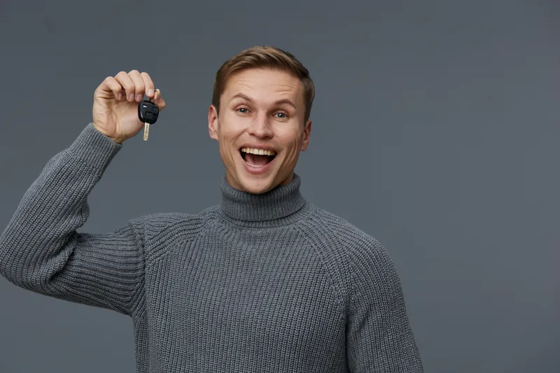 Happy young Caucasian man in gray sweater holding car key presenting with smile isolated on gray background studio portrait. People lifestyle concept