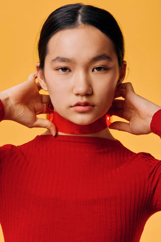 Woman with hair tied back wearing red sweater adjusting red scarf on neck against orange background in studio. Portrait of serious young female with natural makeup and minimal accessories.