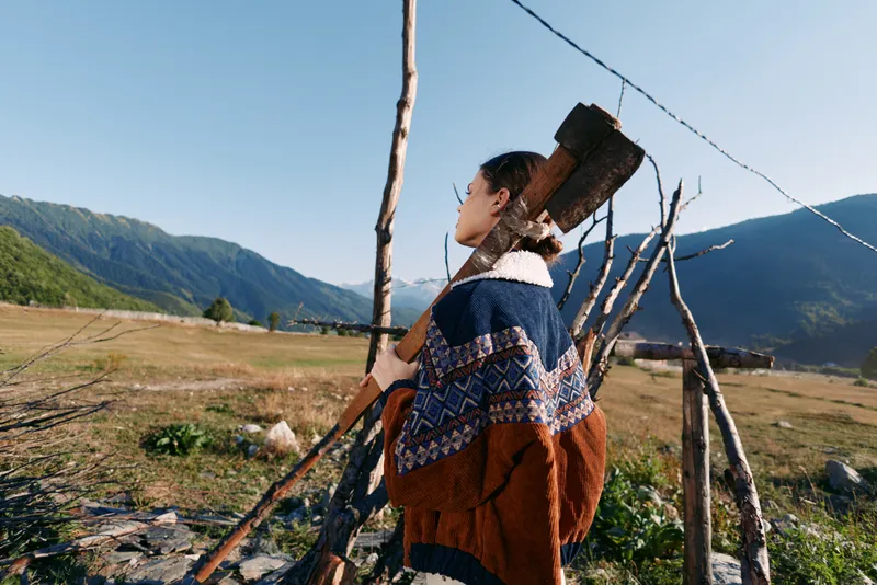 Woman with axe standing in rural countryside field near a wooden fence, wearing patterned sweater and carrying wood for chopping in mountains valley, outdoor rustic lifestyle portrait.