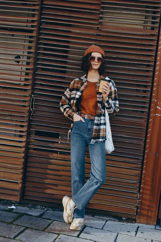 Woman on urban street holding coffee, casual autumn style in beanie and sunglasses, plaid shirt and jeans. Authenticity, candid lifestyle, golden hour glow and mindful living mood.