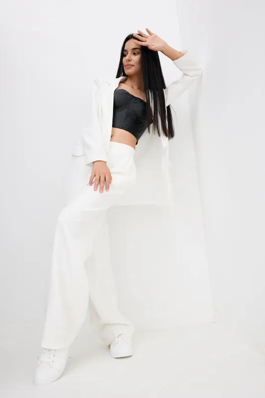 Stylish woman in white pants and blazer with black top posing against a white wall, showing modern fashion and confident attitude in a minimalist setting.