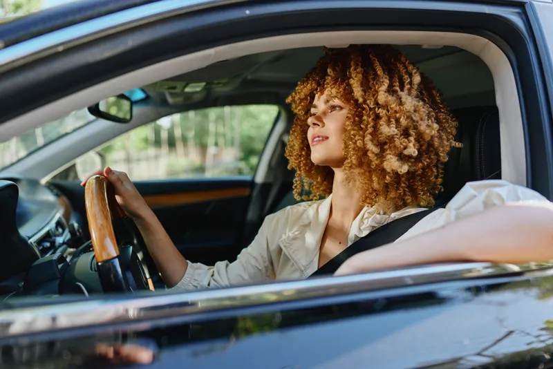 Young woman driving car with curly hair and casual clothing, enjoying calm sunny day and relaxed mood during urban commute through green surroundings.