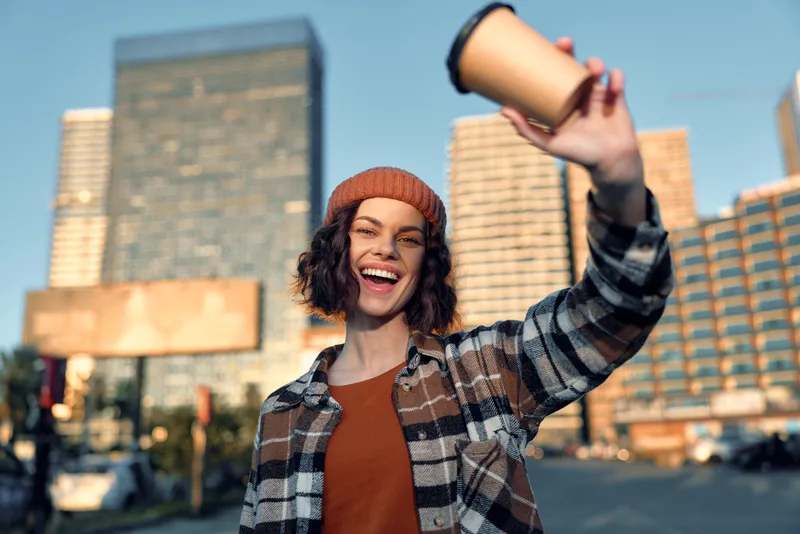 Woman holding coffee cup and smiling on city street at golden hour, urban portrait with sunlight morning vibe. Authenticity, candid lifestyle, golden hour glow, mindful living, emotional storytelling.