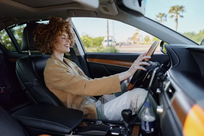 Cheerful woman driving outdoors, smiling with eyes forward, sunlit moment, relaxed posture, retro film color tones, warm palette, car interior details, natural confidence, candid lifestyle
