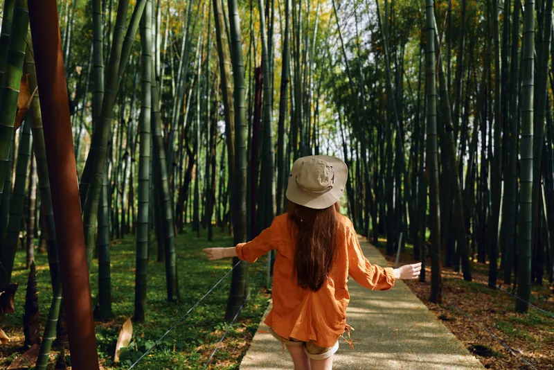 Bamboo woman on forest path wearing beige hat and orange shirt, walking with arms outstretched along shaded boardwalk amid tall bamboo stalks and dappled sunlight.
