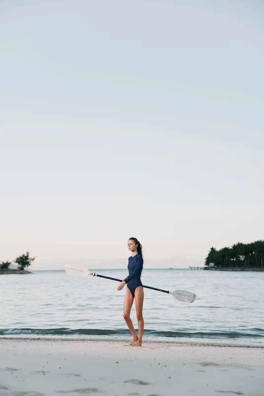 A lone figure stands on a calm sandy shoreline, holding a double paddle across a tranquil sea as soft light washes the horizon and the water reflects pastel sky.