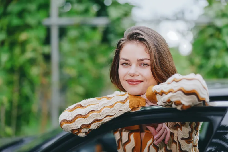 Portrait of a cheerful young woman resting her arms on a car window frame, smiling softly in a leafy outdoor setting.