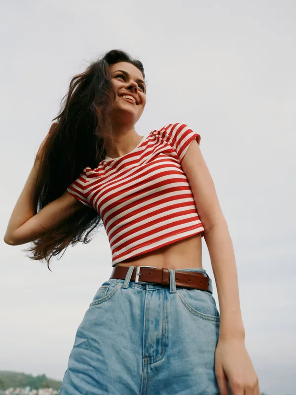 A cheerful young woman poses outdoors in a red and white striped crop top and high waisted denim jeans, showcasing bright style, confidence, and carefree energy under a clear sky.