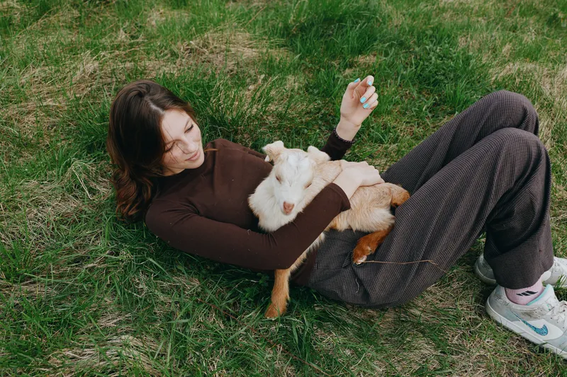 A relaxed woman lies on the grass with a friendly goat kid resting on her chest in a sunny outdoor scene. A lifestyle portrait of bonding, care, and gentle animal interaction.