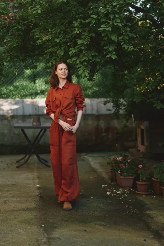 Woman in an orange jumpsuit posing in a sunny garden, a casual lifestyle portrait outdoors, natural backdrop, relaxed fashion mood and warm, approachable expression