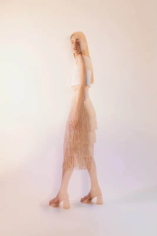Young woman in a white top and beige fringe skirt poses gracefully in soft lighting. Concept of woman beauty fashion with elegant and modern style in a minimal studio setting.