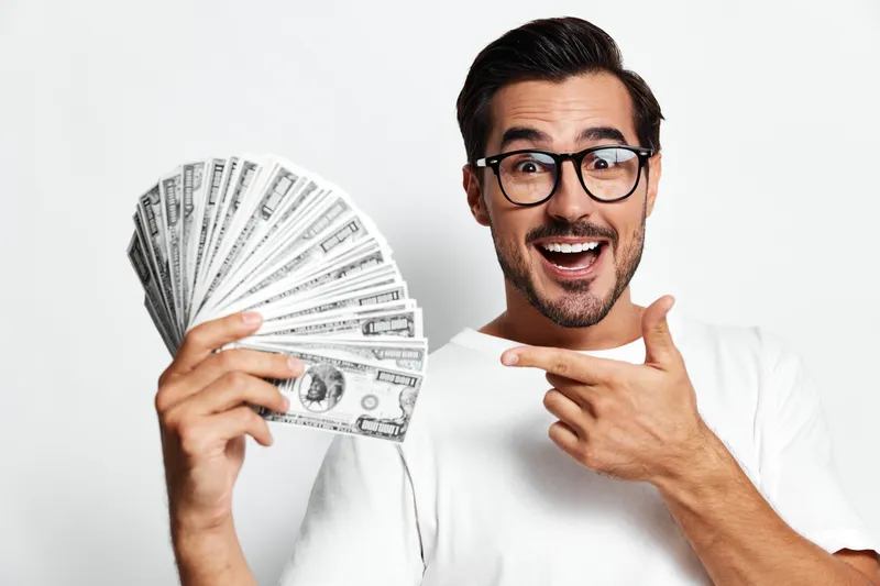 Happy smiling man with glasses holds a fan of cash while pointing at it, showcasing excitement and success, isolated on a plain white background.