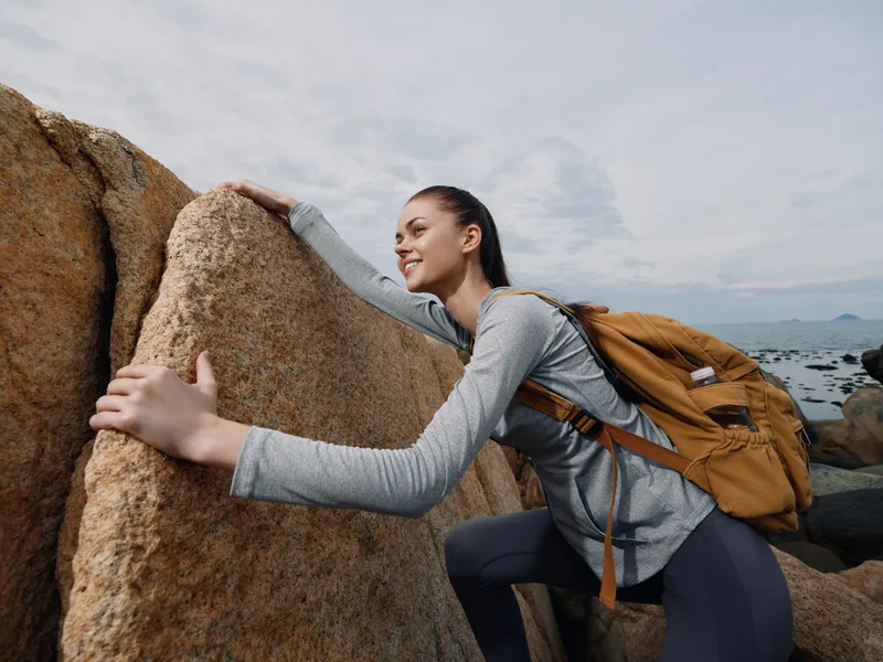 Young female hiker in sportswear climbing rocks, showcasing determination and adventure, with a backpack, against a cloudy backdrop, ideal for outdoor lifestyle concepts