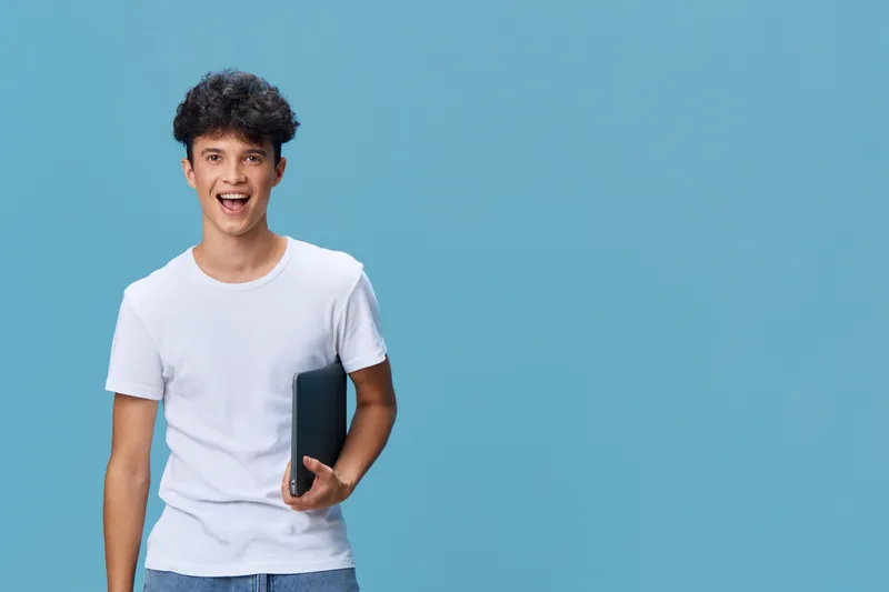 excited young man holding a laptop standing against blue background, wearing casual white t-shirt and jeans, with curly hair and happy expression