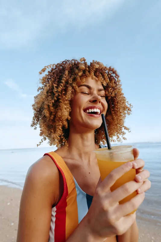 Joyful woman on the beach, smiling with sunlit skin, holding a juice glass with straw, wearing a colorful swimsuit, relaxed pose as waves roll in nearby