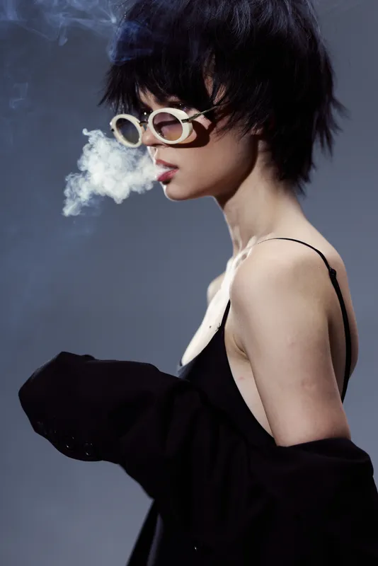 Stylish woman in sunglasses exhaling smoke, wearing black dress and jacket, dark background, modern fashion concept.