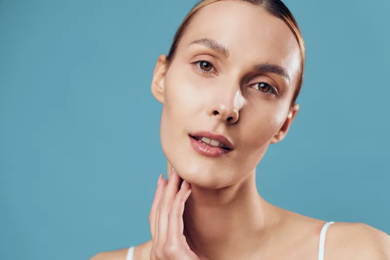 A close-up portrait of a young woman with clear skin and natural makeup against a blue background. She gently touches her neck, highlighting skincare and beauty concepts.
