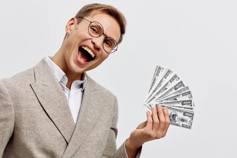 Confident smiling man in beige suit glasses holding fan of dollar bills, expressing happiness and success, isolated on light background. Business success concept, financial achievement, people lifesty