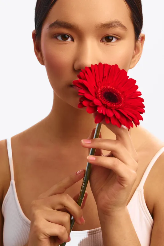 Asian woman holds a bright red flower near her face, smiling softly in a clean studio setting. The composition conveys calm elegance and a gentle, fresh vibe with a minimal aesthetic.