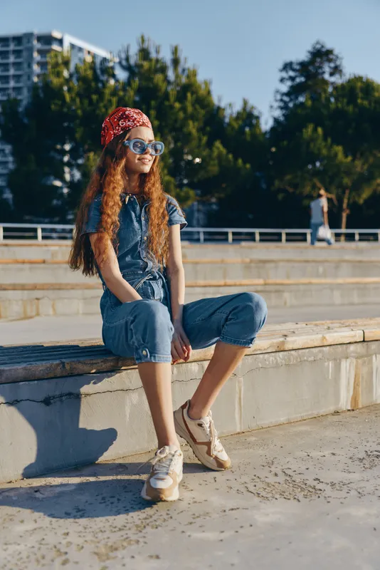 Lifestyle woman in modern boho-western denim street style sits outdoors wearing sunglasses and a red bandana with film color tones for an urban casual look.