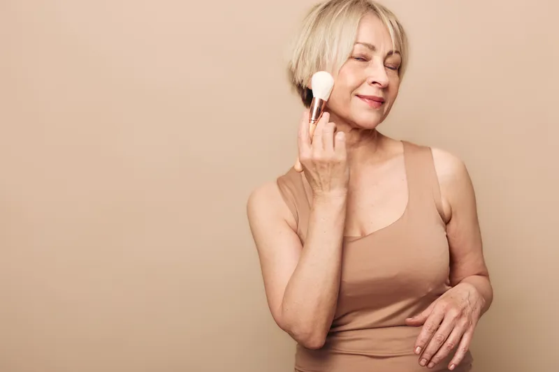 Mature woman with short blonde hair applies makeup powder on face with brush, eyes closed and smiling. Neutral beige background creates soft, calm atmosphere for beauty and skincare concept.