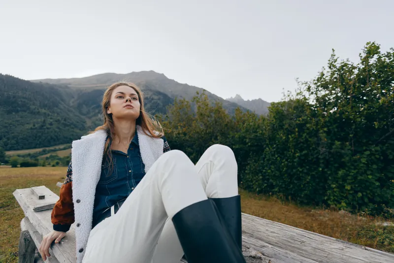 Woman in a coat and boots relaxing on a wooden bench in the countryside with mountains and nature in the background, casual outdoor portrait showing leisure, calm and scenic mood.