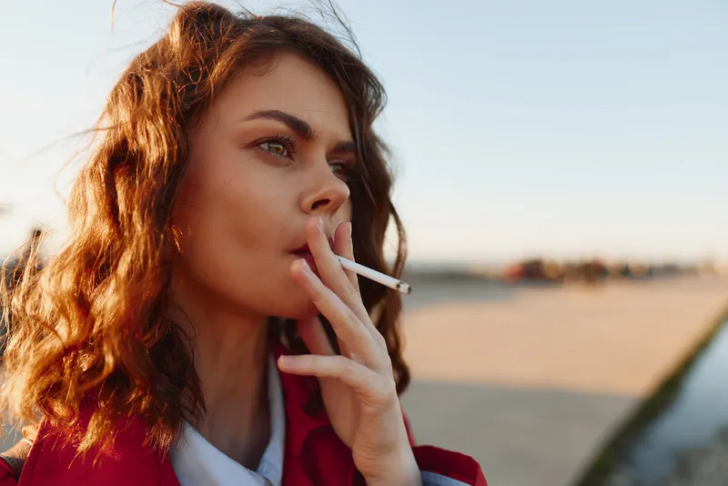 Young woman with curly hair smoking a cigarette outdoors during sunset. Close-up portrait with natural light and blurred background. Concept of addiction and contemplation.