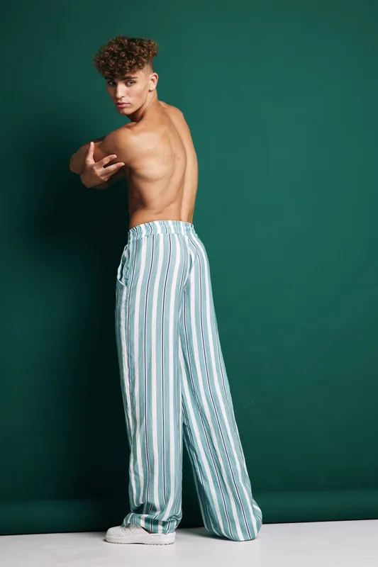 Young man with curly hair posing shirtless in striped wide pants and white sneakers against green background. Fashion model showing back and side profile with confident expression.