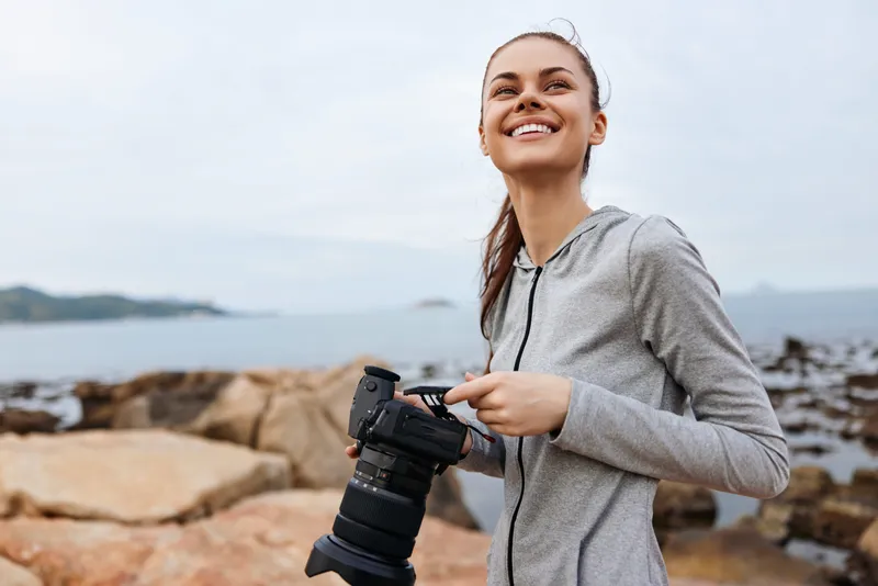 Happy female photographer standing by the ocean with a camera, wearing a casual gray hoodie, capturing beautiful moments at the seaside with a joyful expression