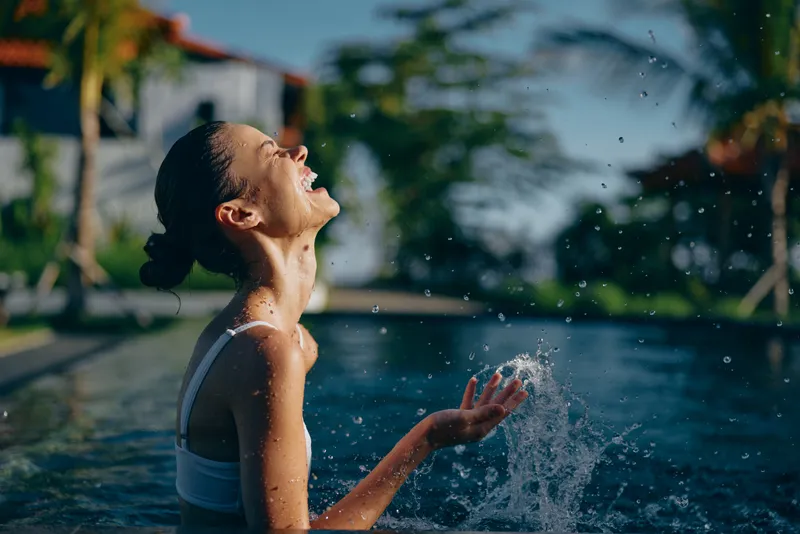 Girl enjoying water in a pool, laughing as spray arcs around her, tropical outdoor scene, summer leisure, carefree mood, bright sunlight, candid moment of joy and outdoor recreation