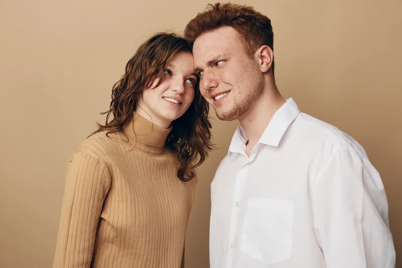 Young couple with casual clothes stands close together on beige background. Woman looks at man with smile while man shows confused expression. Concept of relationship, emotions, and communication.