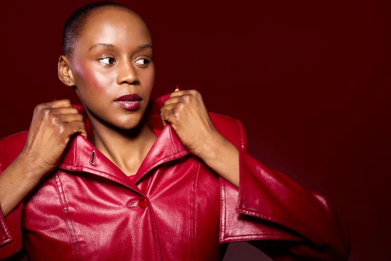 Confident African woman with dark skin adjusts collar of red leather jacket against dark red background. Fashion portrait, style, elegance, and bold color contrast for modern urban look.