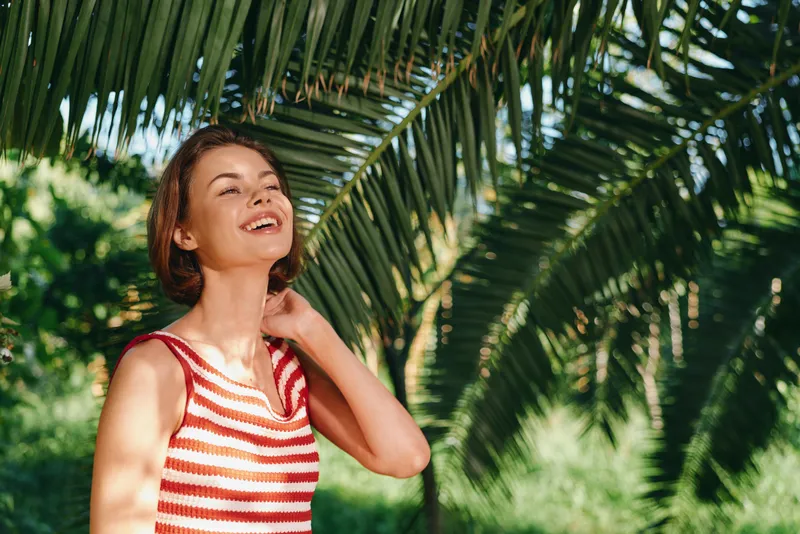 A cheerful woman enjoys an outdoor scene beneath palm fronds. She wears a striped top, smiles warmly, and projects positive energy, creating a vibrant, relaxed summer mood.