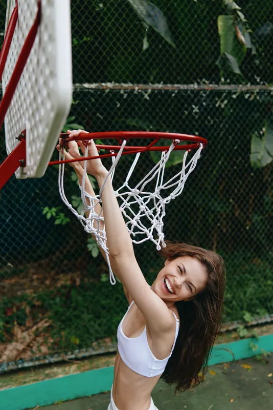 Joyful young woman in a sports bra hanging on a basketball hoop, expressing happiness and energy in a vibrant outdoor setting