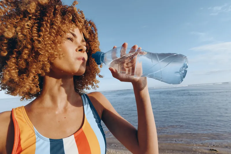 Woman with curly hair drinking water from bottle outdoors on sunny beach, ocean horizon background, summer hydration, relaxed stance, clear blue sky, gentle breeze, soft light