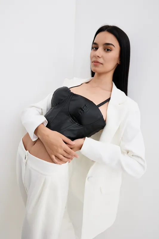 Elegant woman with long black hair wearing a white suit and black corset standing confidently against a plain white background in a minimalist fashion style.