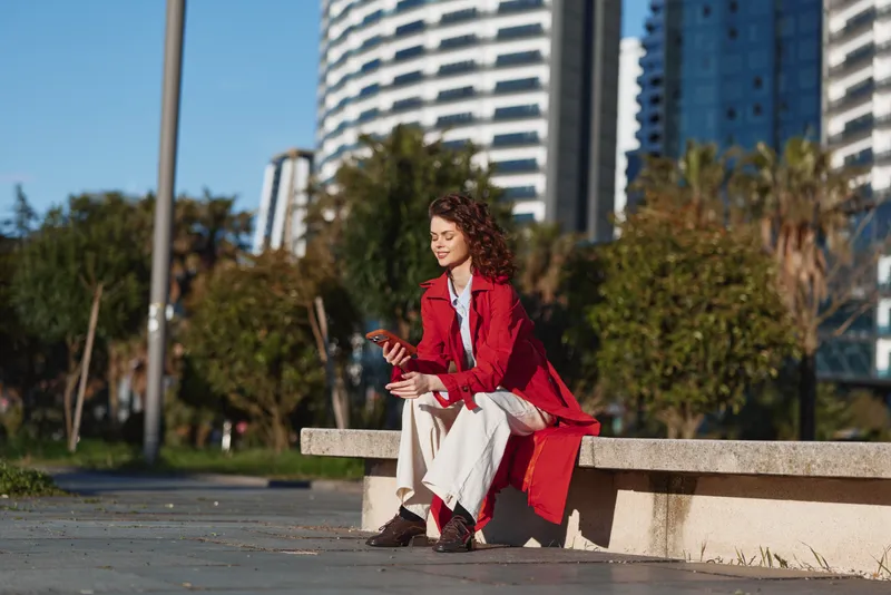 Young woman with curly hair in a red coat sits on a bench outdoors using a smartphone with a relaxed and focused expression in an urban park setting.