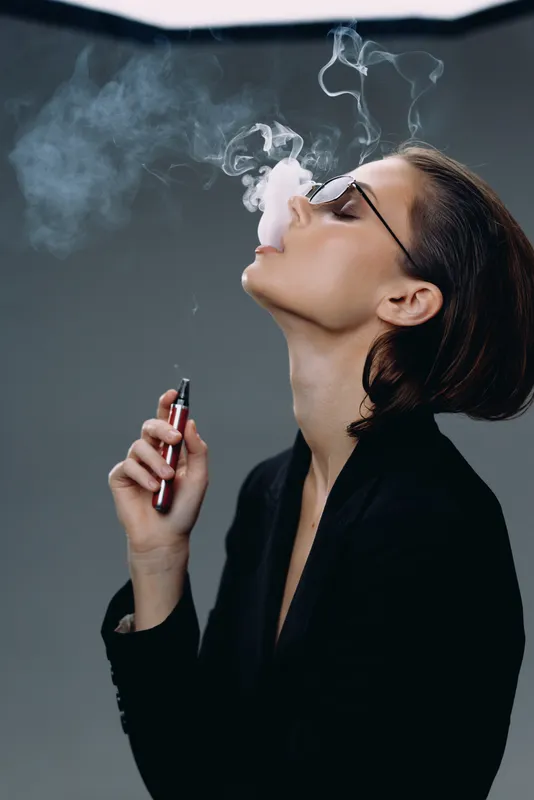 Woman vaping with glasses exhaling smoke in dark background, modern style, relaxing and casual atmosphere.