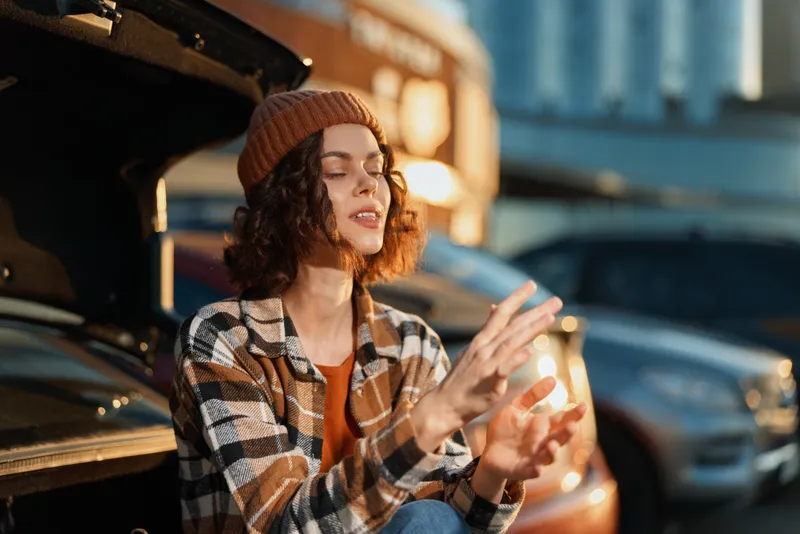 woman car trunk beanie plaid urban relaxation candid young woman sits at open car trunk during golden hour, eyes closed with hands warm in cozy beanie, mindful living and emotional storytelling