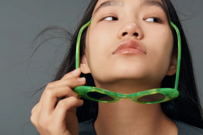 Woman with long black hair holding green sunglasses near face with thoughtful expression on grey background. Beauty, fashion and casual portrait with lifestyle and style concept.