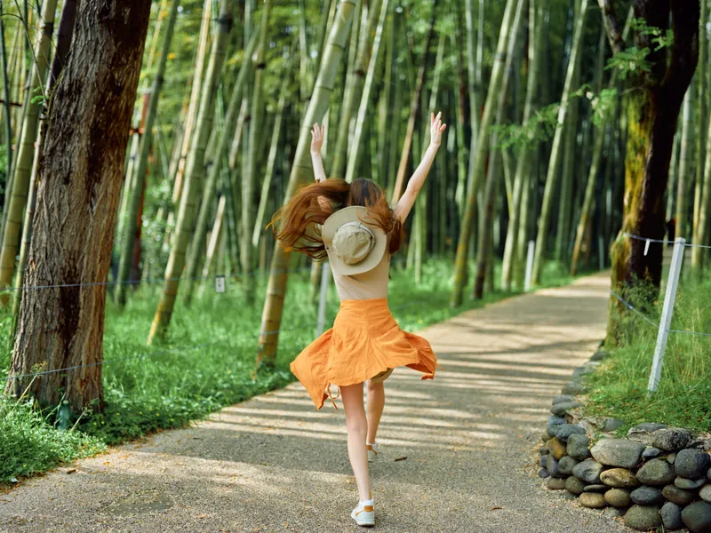 Woman in an orange dress and straw hat runs joyfully along a gravel path through a bamboo forest, back view in summer sunlight, carefree motion and outdoor nature escape.