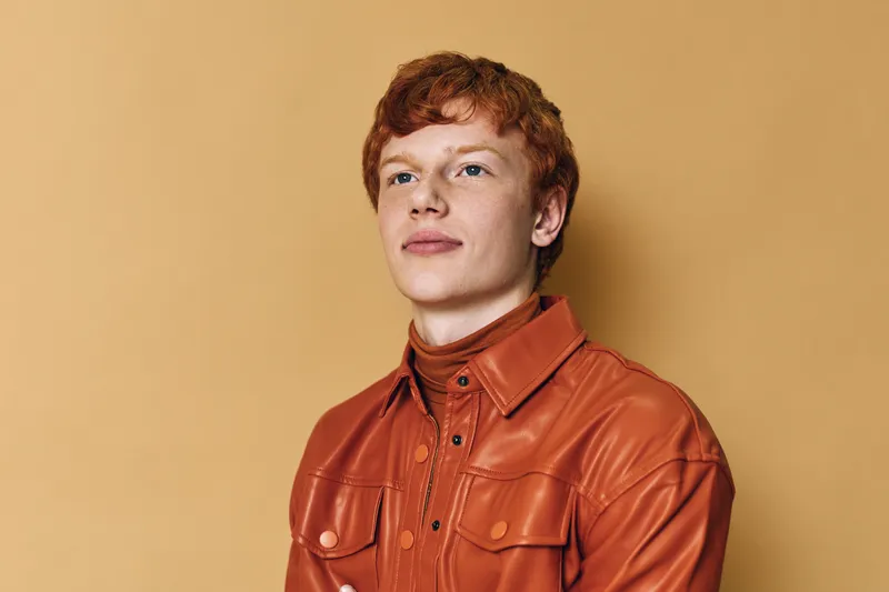 Portrait of young man with red hair wearing orange leather jacket looking away with confident expression on beige background. Studio shot, casual fashion and style.
