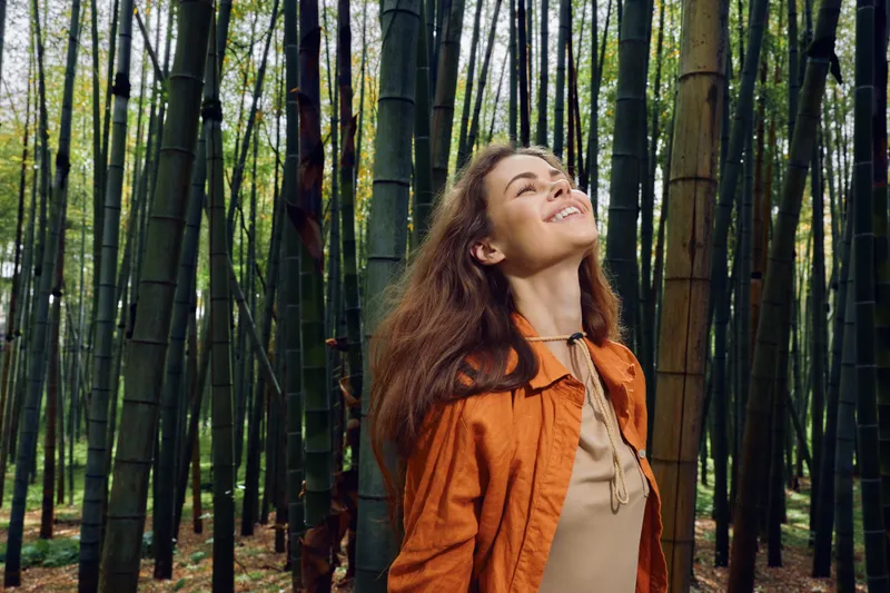 Woman in a bamboo forest smiles while looking up, enjoying nature outdoors with sunlight filtering through tall green stalks; portrait of happy relaxed adult in orange jacket.