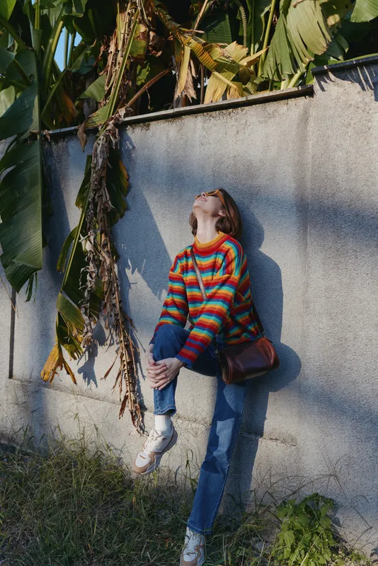 Woman in a striped sweater leaning against a concrete wall in tropical outdoor light, wearing blue jeans, white sneakers and a crossbody bag, smiling in a relaxed travel lifestyle portrait.