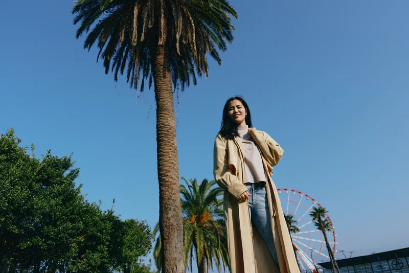 Outdoor scene with a stylish woman standing near tall palm trees and a distant ferris wheel under a clear blue sky, showcasing a casual coat and denim for a relaxed urban summer mood.