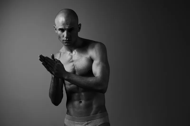 Muscular man in black and white, showcasing strength and determination, poses with clenched hands against a subtle gradient background, highlighting his powerful physique.